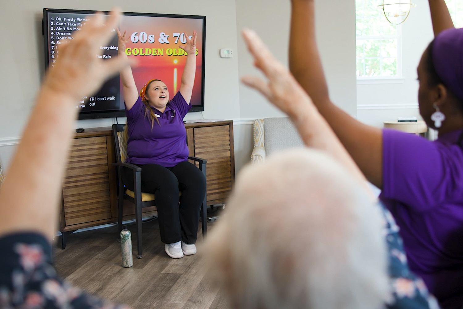 A woman in a purple polo raises her arms from a chair as a group follows along near a screen listing songs from the 60s and 70s at Arbor Terrace Sudley Manor. Senior activities with music and movement add fun to an active senior lifestyle in Manassas, VA.