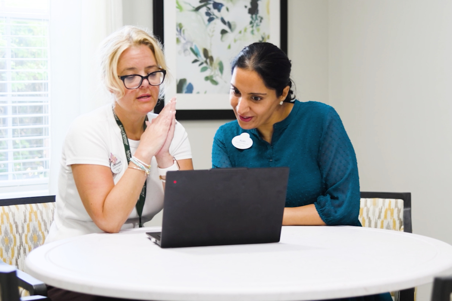 Two women, one in glasses and a white tee and one in a teal blouse, look at a laptop together at a round white table beneath botanical art at Arbor Terrace Sudley Manor. Teamwork shines at this Manassas VA senior living community.