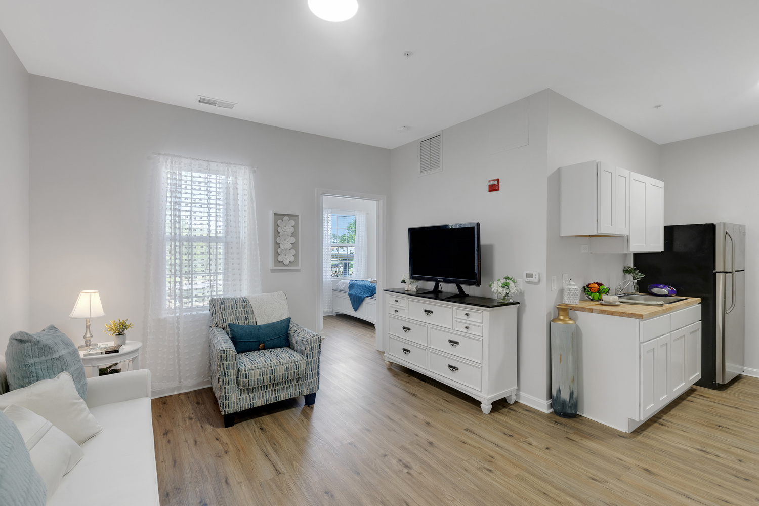 An open apartment at Arbor Terrace Sudley Manor with a sofa, patterned armchair, TV atop a white dresser, kitchenette with a fridge, and a bedroom beyond, showcasing an apartment at this community offering assisted living and memory care in Manassas, VA.