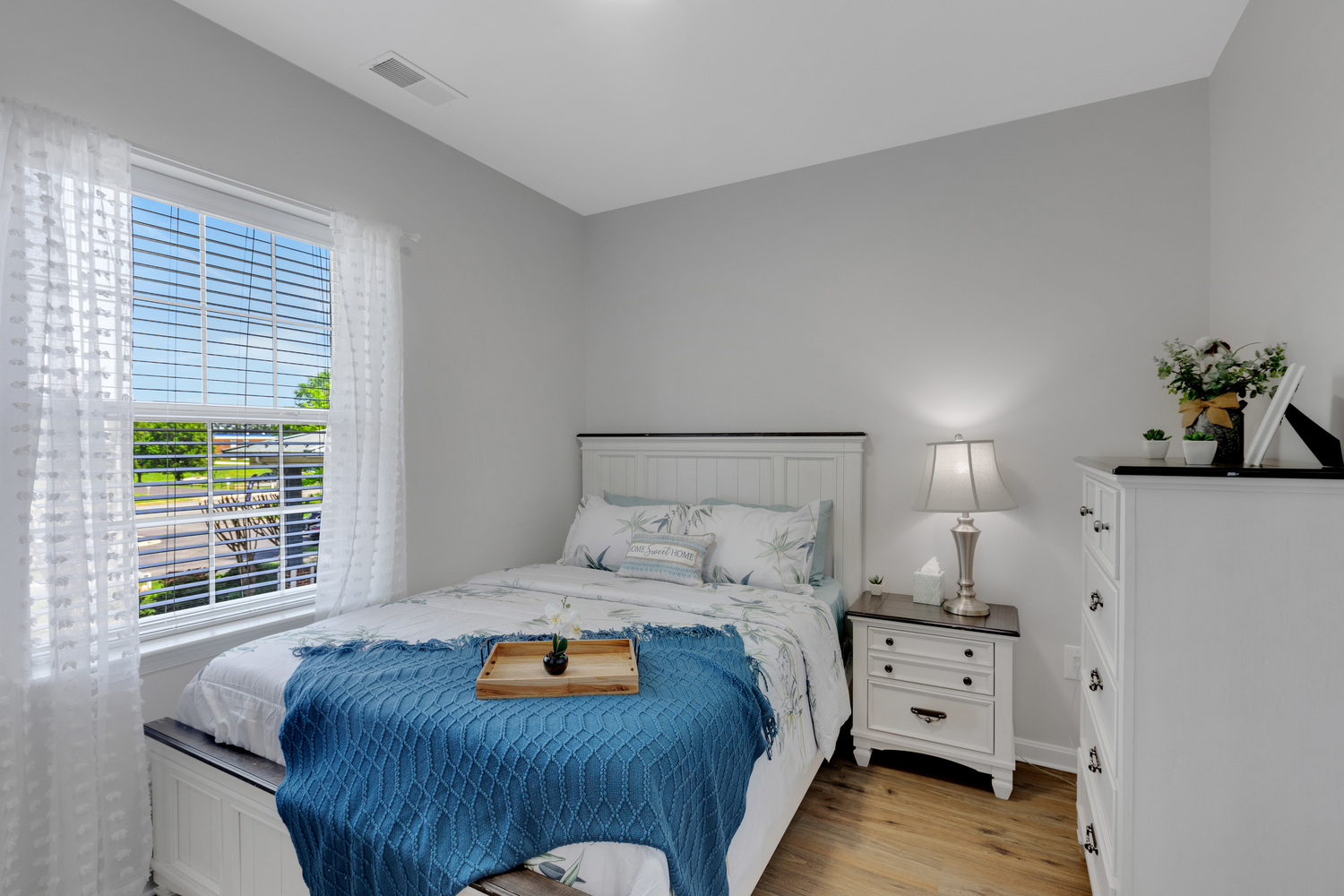 A bedroom in an apartment at Arbor Terrace Sudley Manor with a white bed frame, blue throw blanket, nightstand and lamp, and sheer curtains at a sunny window, showcasing an apartment for assisted living and memory care in Manassas, VA.