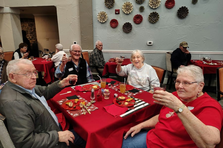 A group of older adults raises glasses in a toast at a festively decorated red-tablecloth dining table — joyful holiday celebrations at senior living Lakeway Texas.