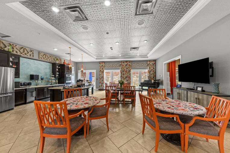 Bistro style dining space at The Vantage at City View with orange chairs at floral topped tables, a pressed tin tray ceiling, pendant lights over the kitchen counter, and a grand piano by the windows, a bright setting for on-site dining at this senior living community in Fort Worth, TX.