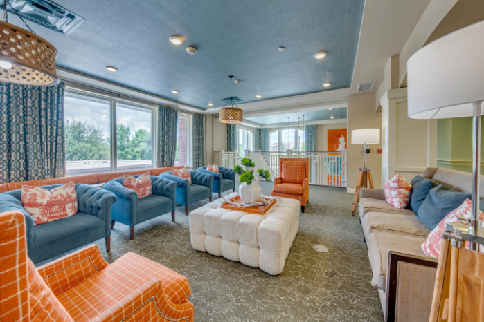 A lounge at The Vantage at City View with blue tufted chairs and orange wing chairs around a large ottoman, tall windows framing treetops, and a balcony overlook, a bright gathering space at this senior living community in Fort Worth, TX.