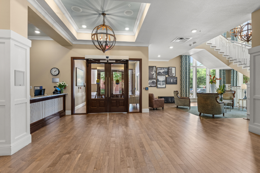 Lobby at The Vantage at City View looking toward the wood double entry doors, with an orb chandelier in the tray ceiling, the front desk and wall clock at left, framed black and white photos, and patterned armchairs below the staircase, senior living in Fort Worth, TX near Cityview.