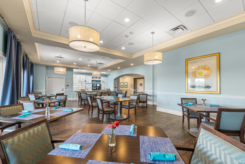 Dining room in the memory care neighborhood at The Vantage at City View with blue placemats, teal napkins, red flowers on the tables, drum pendant lights, and sunburst artwork, showcasing on-site dining and memory care in Fort Worth, TX.