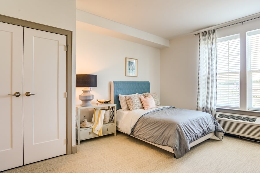 A bright, elegantly furnished resident bedroom featuring a blue upholstered bed, stylish nightstand, and large windows at our assisted living community in Gambrills, MD.