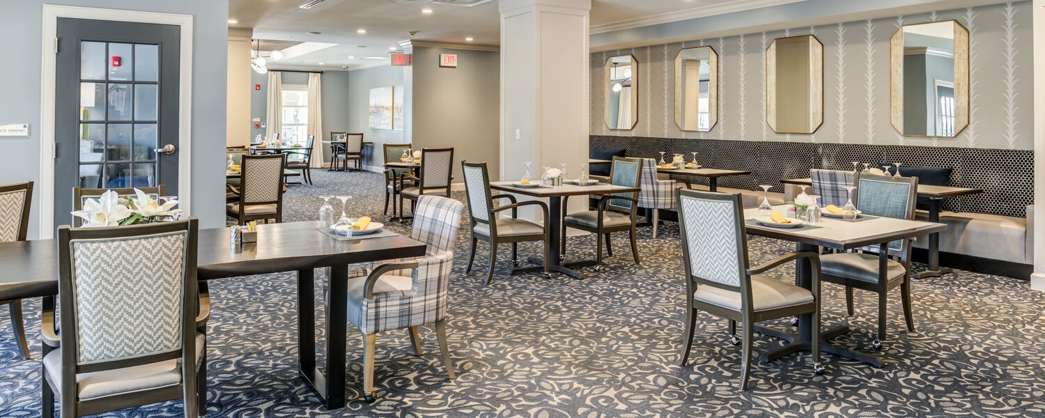 Elegantly set dining tables with patterned upholstered chairs, booth seating, and decorative mirrors in the dining room at Arbor Terrace Willistown, a welcoming assisted living community in West Chester, PA.