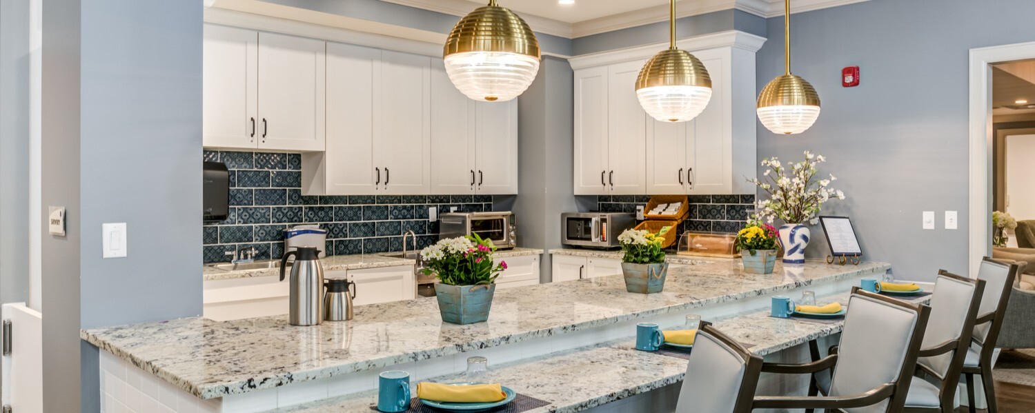 A chef-inspired community kitchen and dining area at Arbor Terrace Waugh Chapel featuring pendant lighting, granite countertops, and elegant place settings, showcasing the resort-style assisted living amenities offered near Waugh Chapel Towne Centre.
