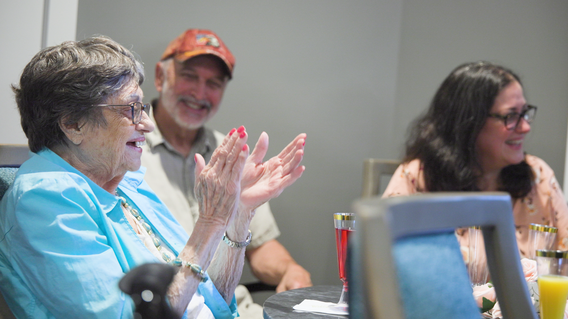 Senior lady in blue clapping while sitting with an older gentleman wearing a red cap and a woman with glasses