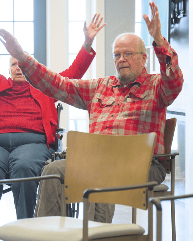 Two residents raise their hands joyfully during a group activity or exercise class, capturing the vibrant, engaging lifestyle at this senior living community in Gambrills, MD.