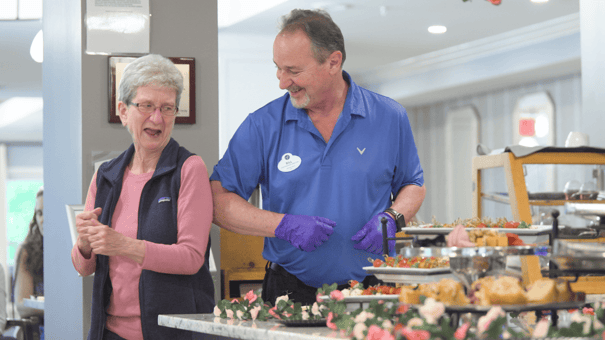 A resident laughs with a staff member beside a beautifully displayed food spread, showcasing the warm dining experience at our resort-style assisted living community in Gambrills, MD.