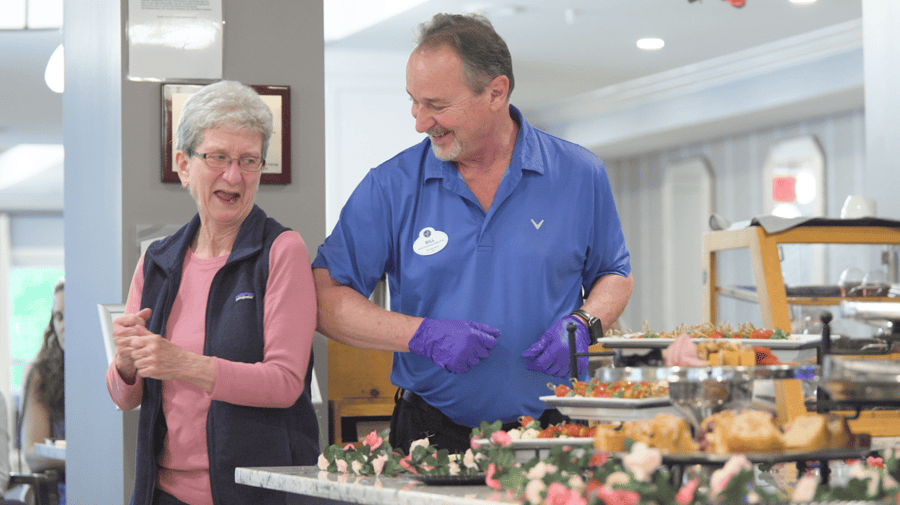 A resident laughs with a staff member beside a beautifully displayed food spread, showcasing the warm dining experience at our resort-style assisted living community in Gambrills, MD.