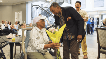 A resident is surprised with a bouquet of flowers during a lively community event, with residents and staff gathered and celebrating in the background at our senior living community in Gambrills, MD.