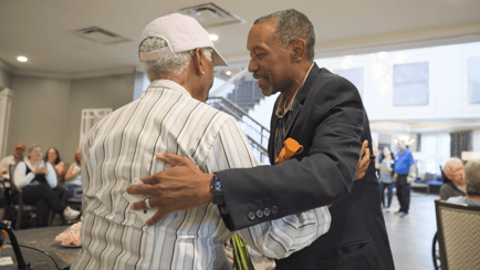 Two people share a warm embrace during a lively community event, with residents gathered and applauding in the background at our senior living community in Gambrills, MD.
