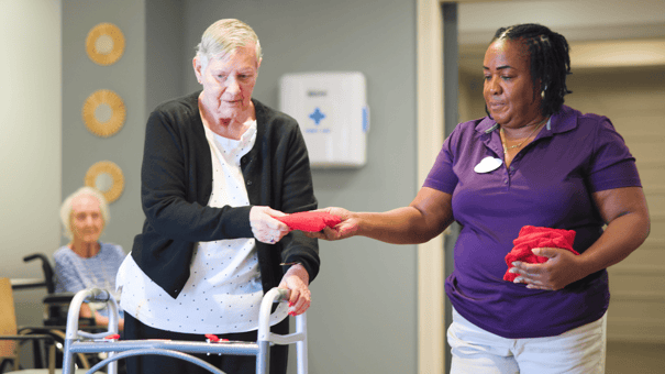 A caregiver in a purple shirt assists an elderly woman using a walker, handing her a red object in an indoor care facility setting.