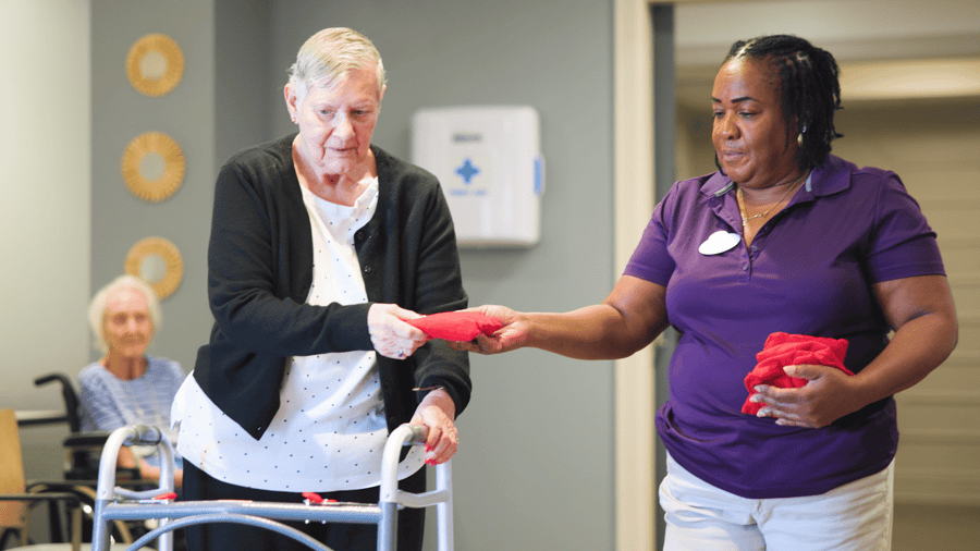 A caregiver in a purple shirt assists an elderly woman using a walker, handing her a red object in an indoor care facility setting.