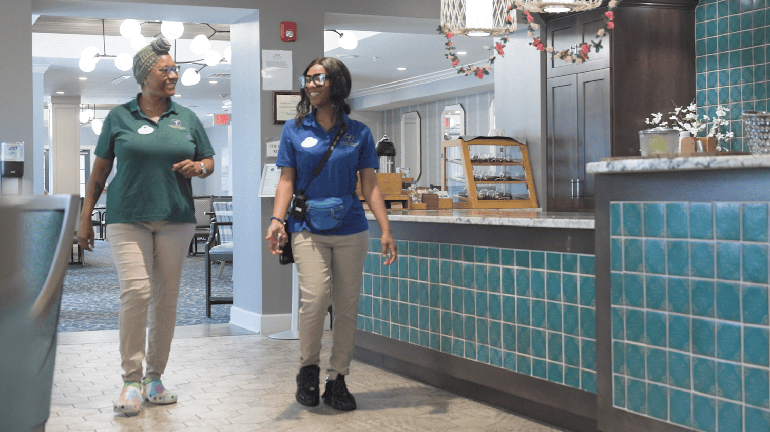 Two staff members walk and converse cheerfully past the teal-tiled front desk in the bright, welcoming lobby of Arbor Terrace Waugh Chapel, reflecting the dedicated and friendly care team that supports residents at this assisted living community in Gambrills, MD.