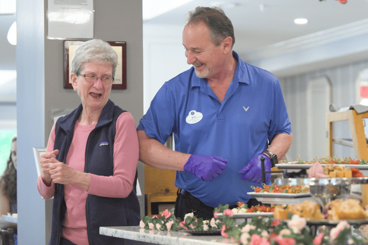 A resident laughs with a staff member beside a beautifully displayed food spread, showcasing the warm dining experience at our resort-style assisted living community in Gambrills, MD.