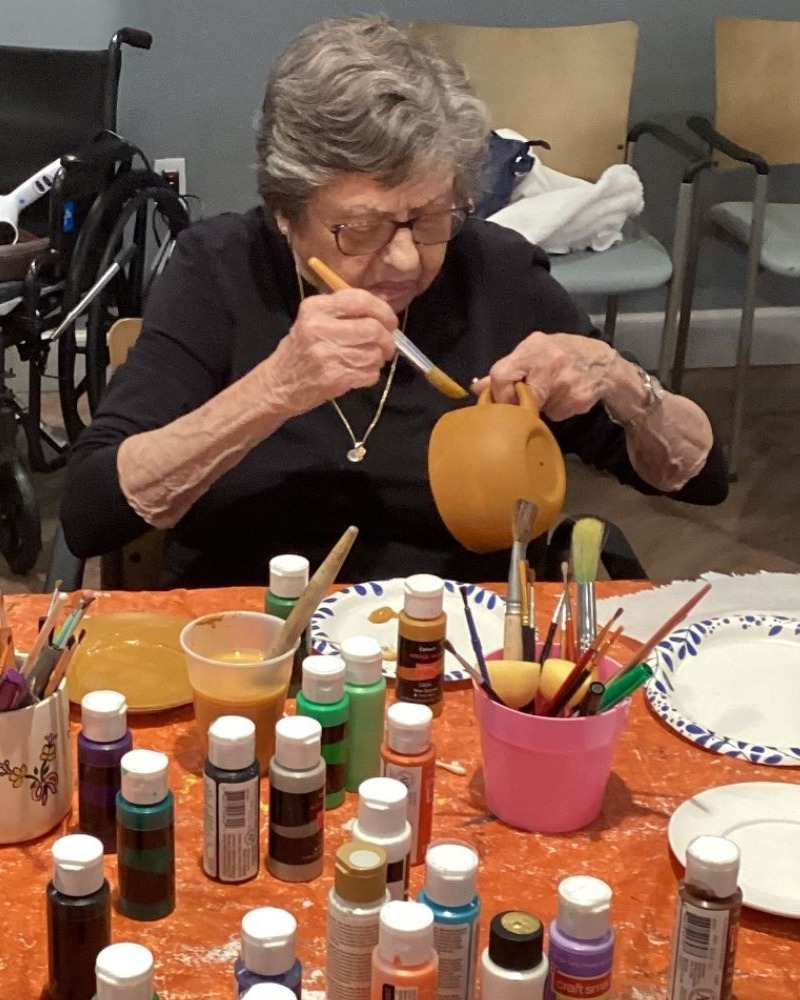 An older adult focuses intently on painting a small pumpkin at a craft table filled with colorful paint bottles and brushes, enjoying a creative activity at this assisted living community in Gambrills, MD.