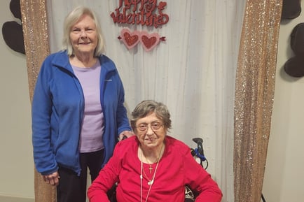 Two residents pose together in front of a festive Valentine's Day backdrop with red hearts and gold sequin curtains at our senior living community in Gambrills, MD.