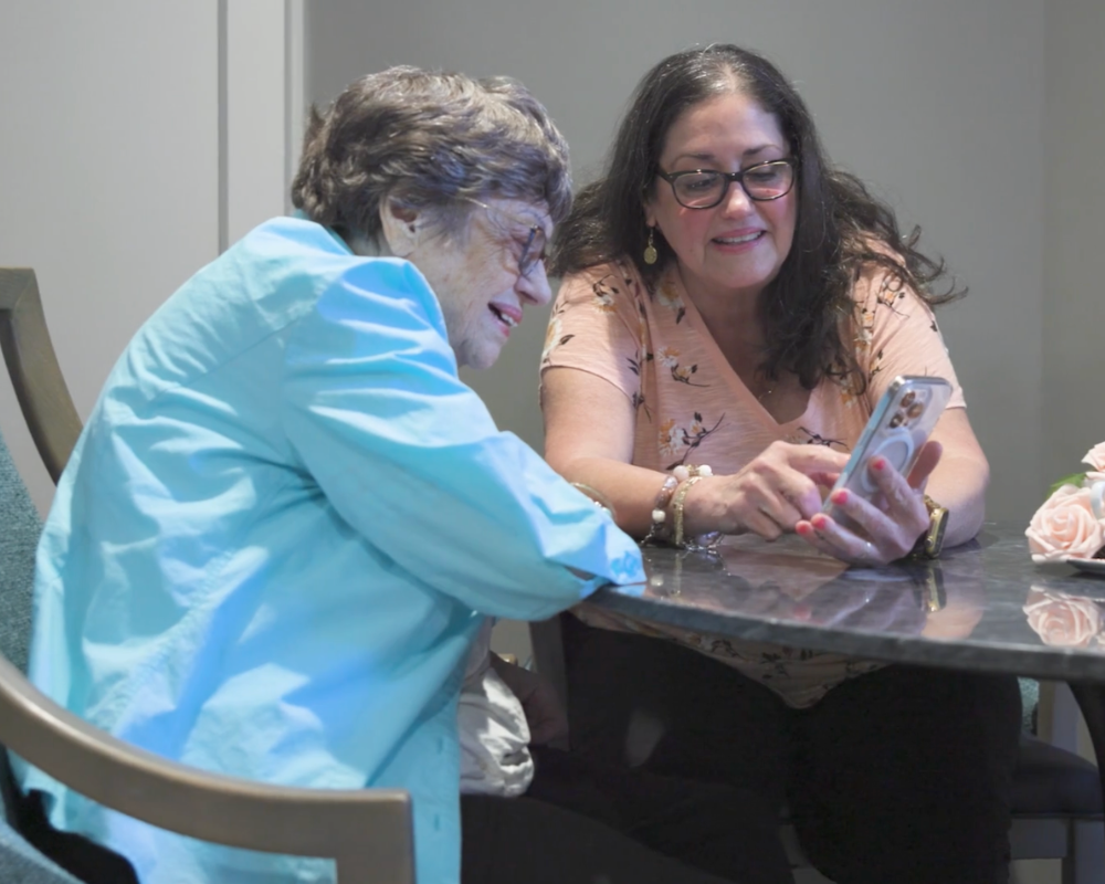 A resident and a visitor look together at a phone, sharing a joyful and intimate moment at Arbor Terrace Waugh Chapel, capturing the warm family connections and supportive community environment at this senior living community in Gambrills, MD.