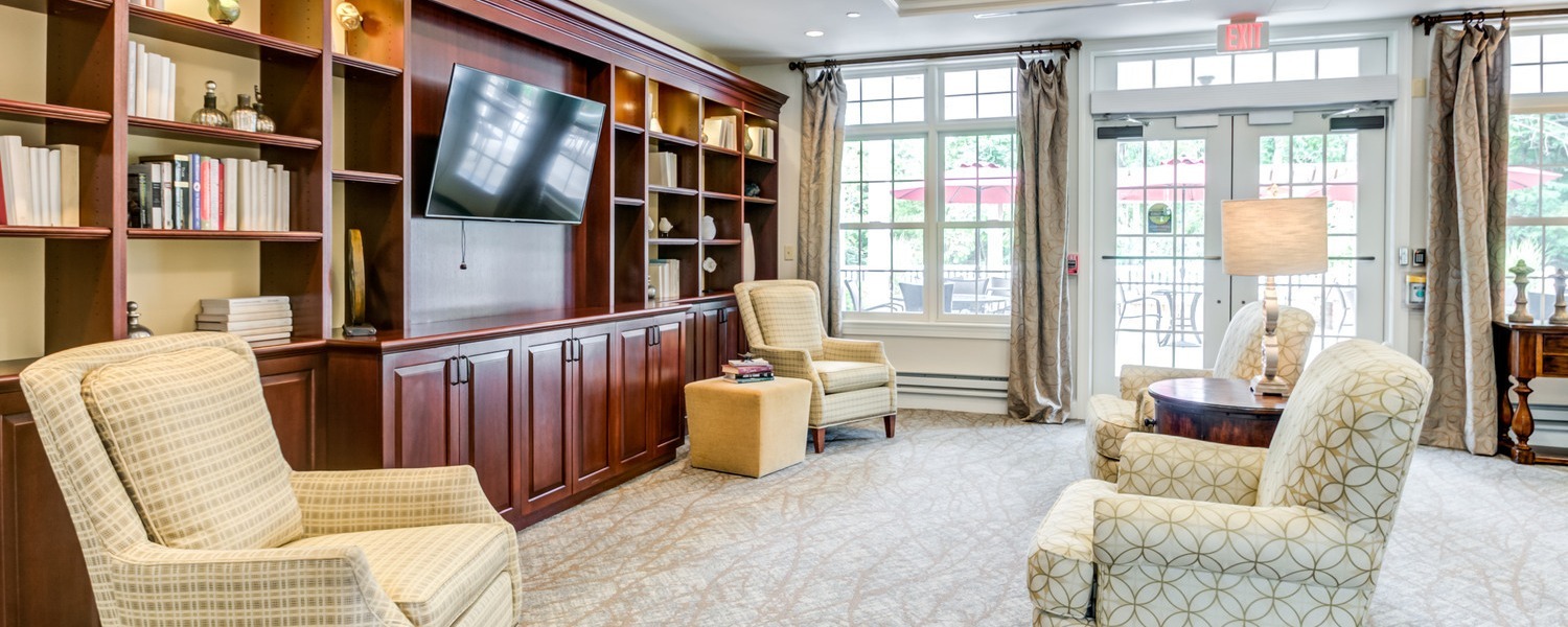 A bright and welcoming sitting room with plush upholstered armchairs, a built-in wood bookcase, wall-mounted TV, and large windows at Arbor Terrace Willistown, a warm assisted living community in West Chester, PA.