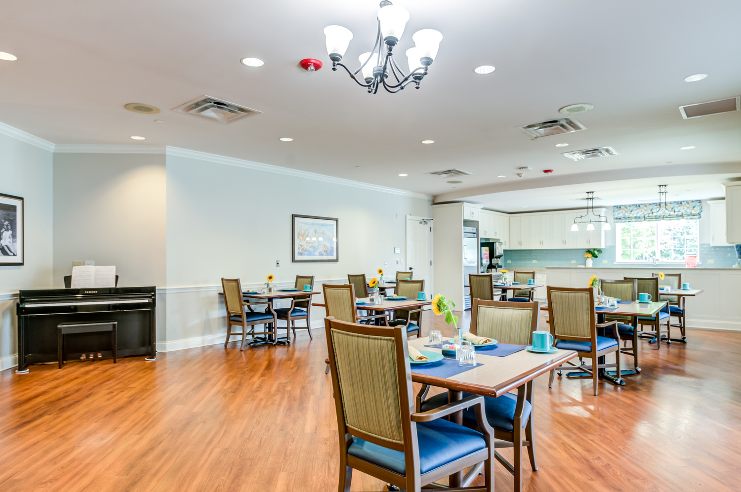 A bright dining and activity room with a piano, sunflower centerpieces, and an open kitchen at Arbor Terrace Willistown, a retirement community in Chester County.