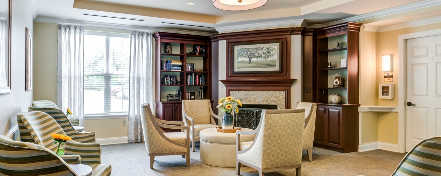 Comfortable resident lounge at Arbor Terrace Willistown in West Chester, PA, featuring cozy armchairs, built-in bookshelves, and a fireplace seating area designed for relaxation and social connection.