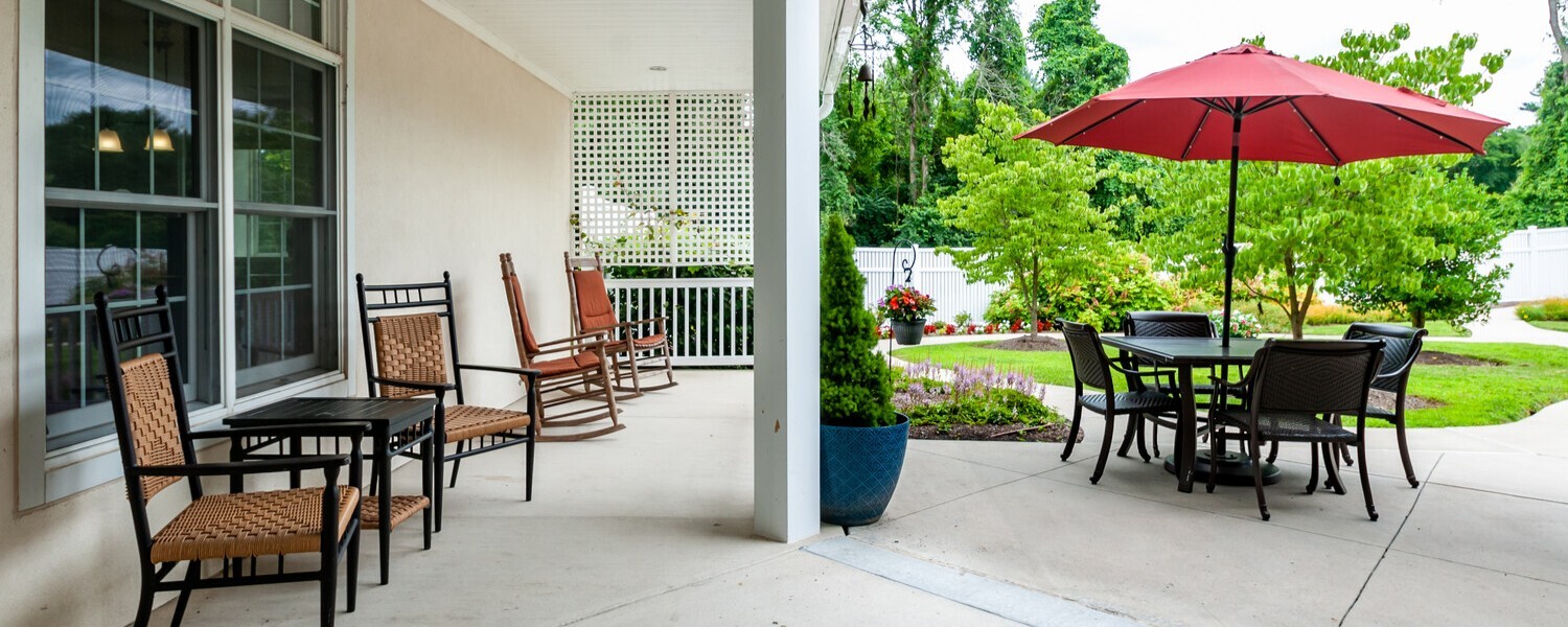 Covered outdoor porch and patio at Arbor Terrace Willistown in West Chester, PA, featuring rocking chairs, shaded seating, and a red umbrella table overlooking landscaped gardens for residents to relax and gather.