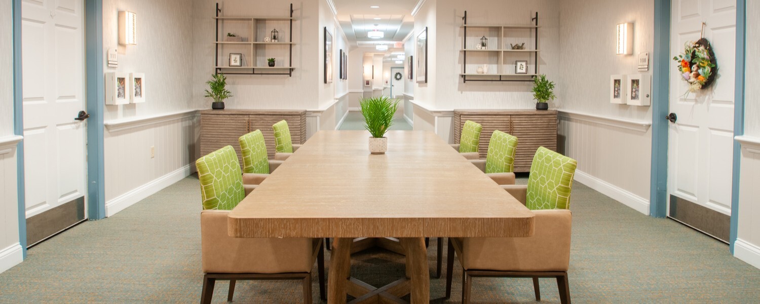 Bright hallway gathering space at Arbor Terrace Willistown in West Chester, PA, featuring a long wooden table with green accent chairs designed for resident activities and social engagement.