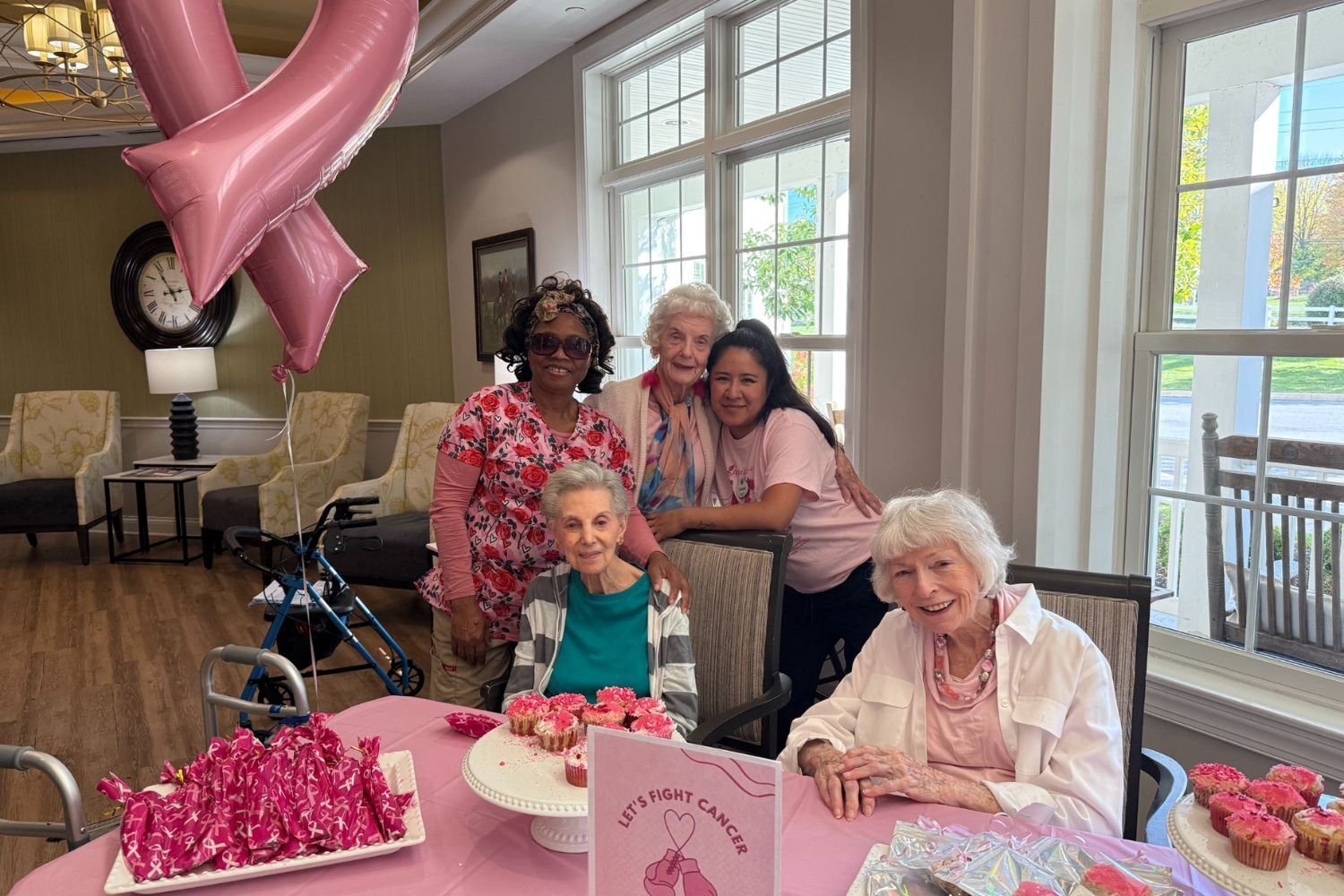 five women gather around a pink table covered in baked goods and smile. some women are standing and some are sitting, most are wearing pink. there is a breast cancer balloon in the room
