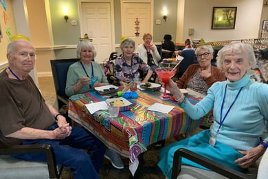 residents smile and sit around a table decorated with a colorful tablecloth. one woman, dressed in all blue, lifts her margarita glass