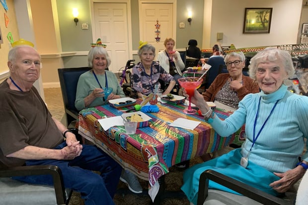 residents smile and sit around a table decorated with a colorful tablecloth. one woman, dressed in all blue, lifts her margarita glass