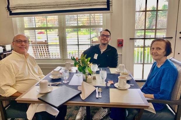 a resident (woman) and two guests (men) sit and smile at a table. They are drinking coffee and water. Both men wear glasses
