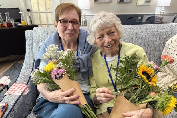 two residents smile and sit on a couch. the women are holding bouquets of flowers wrapped in brown paper