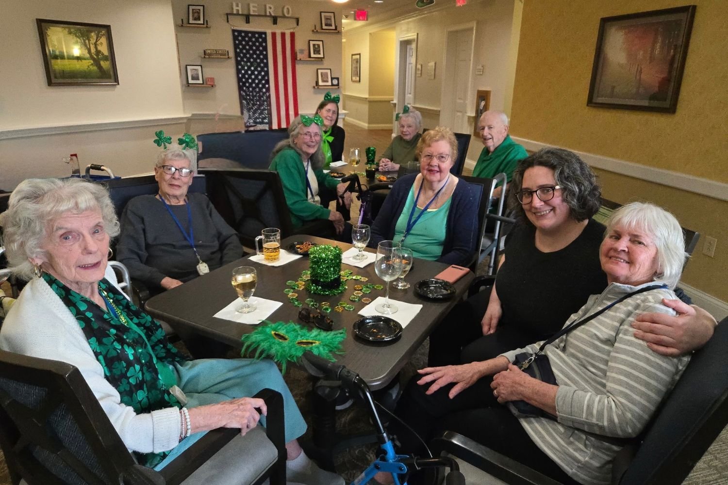 residents smile and sit around a table. the table has St.Patrick's day decor on it and the residents are drinking beverages