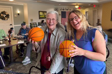a resident and employee smile and stand next to each other. both women are holding small basketballs and standing in a common area with other residents sitting behind them.
