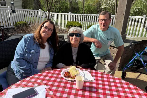 a resident sits outside at a table with her family. There is a plate of food on the table. The sun is out and the resident is wearing glasses.