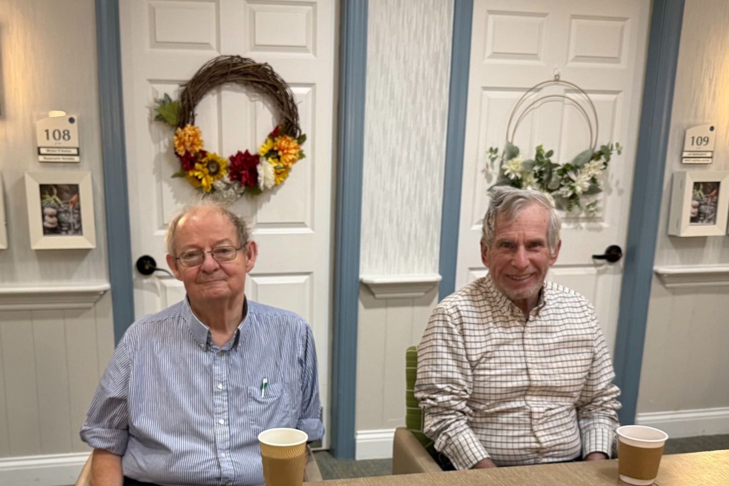 two men sit at a table and smile. they sit in front of two doors with wreaths on them. the residents are wearing button down shirts and have to-go coffee cups in front of them.