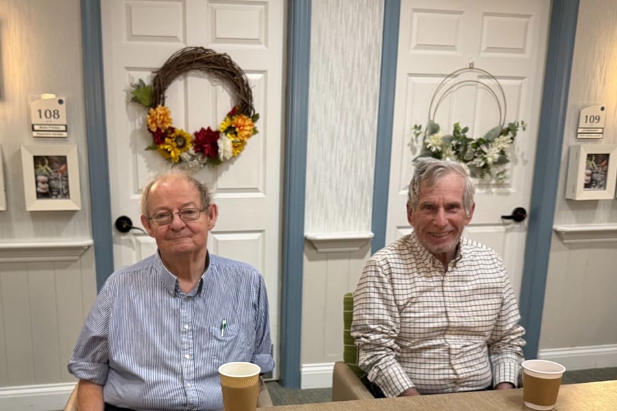 wo elderly men sitting together indoors, smiling, with a decorative wreath on a door in the background.