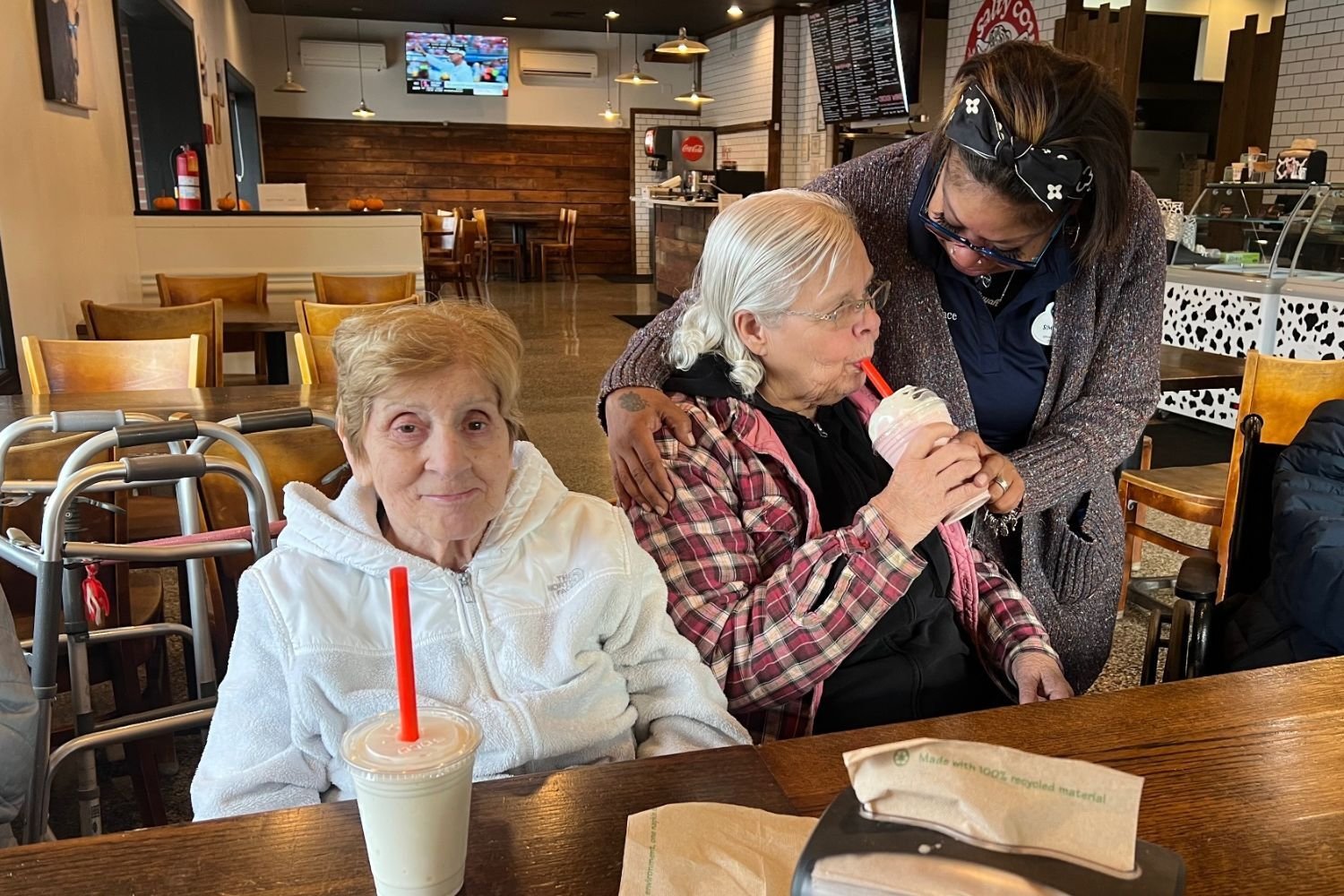 two residents sit at a table with milkshakes. One resident smiles while the other sips her milkshake. An employee puts her hands on the shoulders of the resident sipping the milkshake