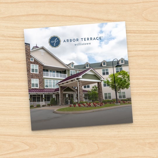 Square photo of Arbor Terrace Williston multi-story building exterior with stone-columned entrance and circular driveway, displayed on a printed card against a wood-grain background.
