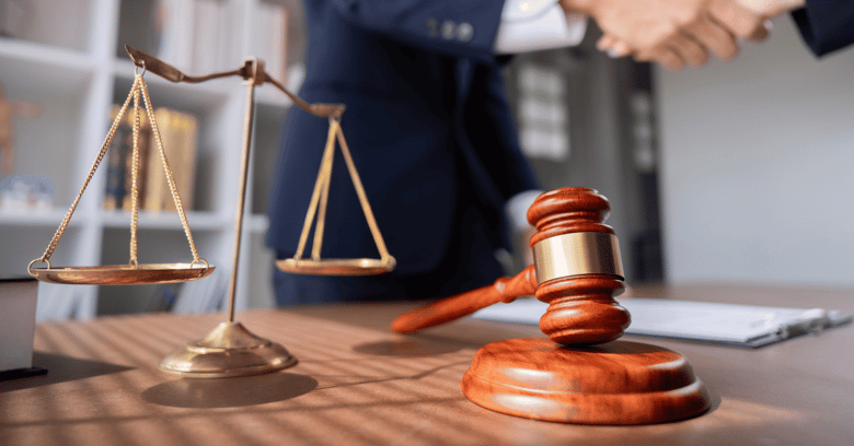 Close-up of a lawyer's desk with a brass scale of justice and a wooden gavel in the foreground, while two people shake hands in the blurred background.