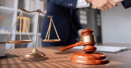 Close-up of a lawyer's desk with a brass scale of justice and a wooden gavel in the foreground, while two people shake hands in the blurred background.
