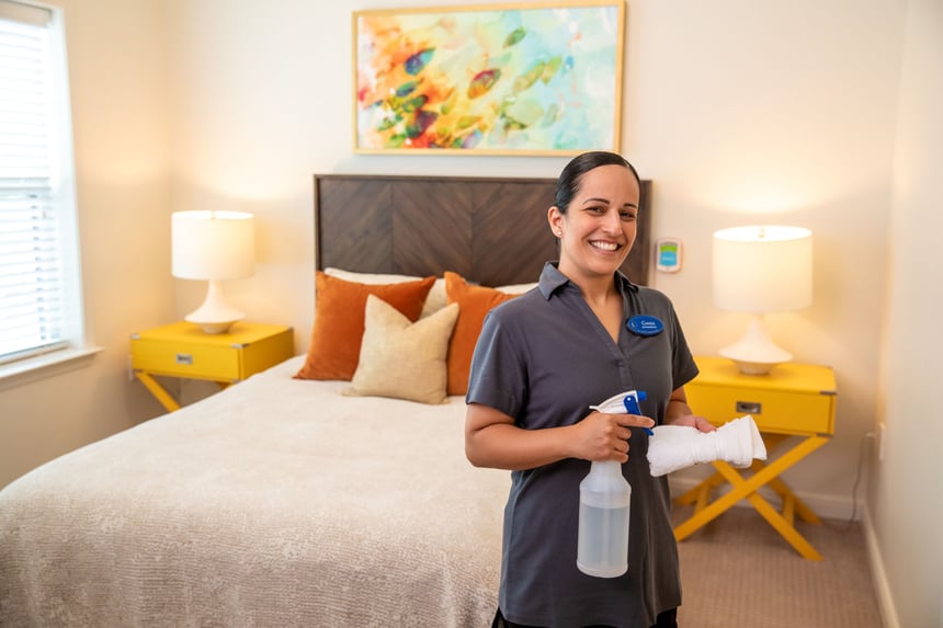 staff member holding a rag and spray bottle standing in front of bed with two yellow night stands in the background