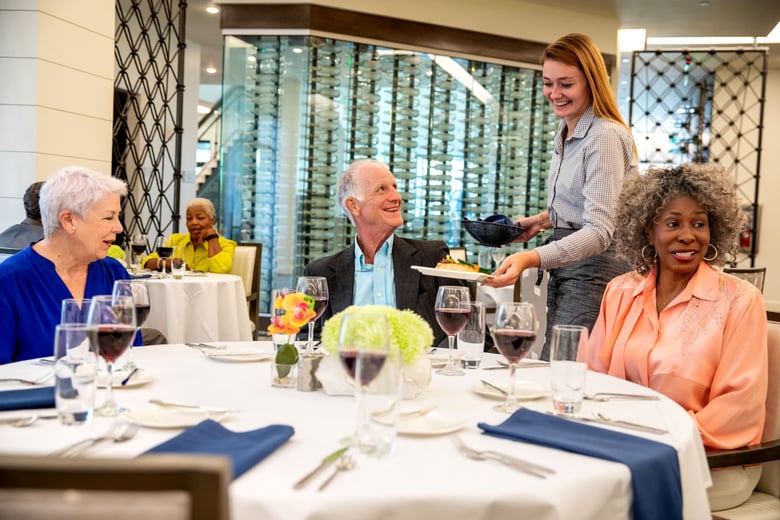 Residents dining at a round table with a white table cloth and blue napkins. server dropping off food at table while three residents are smilling. red wine on the table.