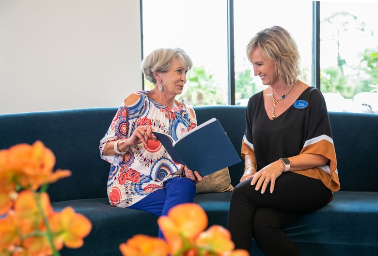 An older woman with gray hair, wearing a colorful patterned top, sits on a teal couch smiling and talking with a staff member wearing a name badge, while looking together at an open book; blurred orange flowers are visible in the foreground.