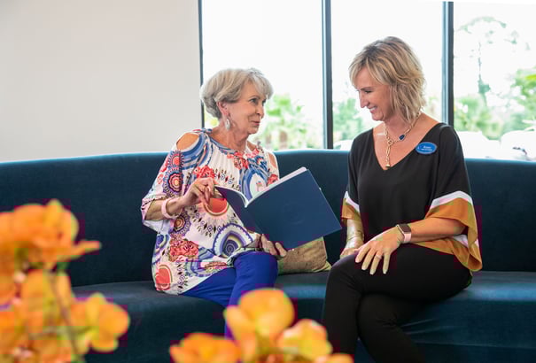 A resident and a staff member sit together on a cozy sofa, reviewing a book in a bright, welcoming common area at our assisted living community in Gambrills, MD.