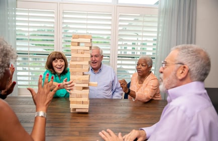 A group of residents laughs and reaches for wooden blocks during a Jenga game around a table at Arbor Terrace Highland Park, with bright shuttered windows behind them. This playful gathering reflects the active senior lifestyle central to senior living in Highland Park, IL.