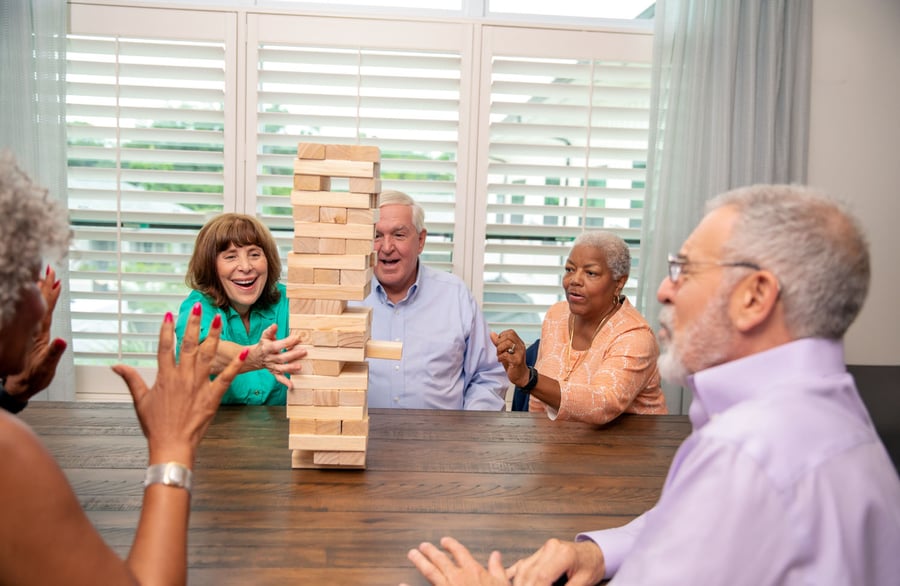 A group of residents laughs and reaches for wooden blocks during a Jenga game around a table at Arbor Terrace Highland Park, with bright shuttered windows behind them. This playful gathering reflects the active senior lifestyle central to senior living in Highland Park, IL.
